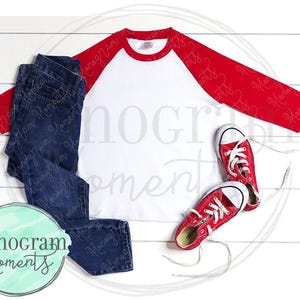 May include: A white and red baseball tee shirt with a red sleeve and a white sleeve. The shirt is laying flat on a white wooden surface with a pair of blue jeans and red sneakers.