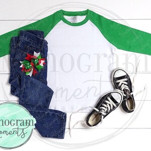 May include: A white and green baseball tee shirt with a pair of blue jeans and black and white sneakers. The jeans have a red and green bow on the front.