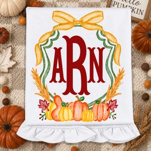 May include: A white ruffled blanket featuring a central red monogram with the letters ARN. The design is framed by a yellow ribbon bow and green laurel branches, with a base of orange pumpkins, gourds, and red autumn leaves.