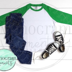 May include: A white and green baseball tee shirt with a white background and green sleeves. The shirt is laying flat on a white wooden surface with a pair of black and white sneakers and blue jeans.