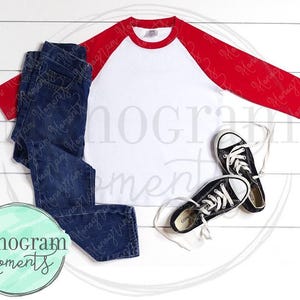 May include: A white and red baseball tee shirt with a white crew neck and red sleeves. The shirt is laying flat on a white wooden surface with a pair of blue jeans and black sneakers.