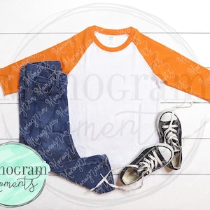 May include: A white and orange baseball tee shirt with a blue denim pair of jeans and black and white sneakers. The shirt has a white body and orange sleeves.