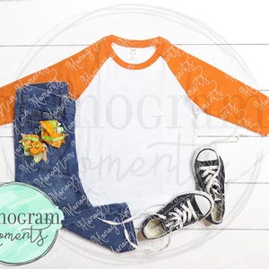 May include: A white and orange baseball tee shirt with a white collar and orange sleeves. The shirt is laying on a white wooden surface with a pair of blue jeans and black sneakers.