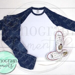 May include: A white baseball tee with navy blue sleeves and a navy blue trim around the neck. The tee is laying flat on a white wooden surface with a pair of blue jeans and white sneakers.