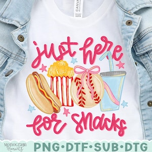 May include: White t-shirt with a colorful graphic design featuring a hot dog, popcorn, baseball, and a drink. The text "just here for snacks" is written in pink script. The shirt is displayed on a denim jacket.