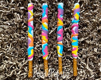 May include: Four pretzel rods dipped in white chocolate and decorated with colorful swirls of blue, yellow, pink, and purple. The pretzel rods are arranged in a row on a brown paper background. The text "Doodle Pops Co" is visible in the lower center of the image.