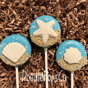 May include: Three blue cake pops with white seashell and starfish decorations. The pops are decorated with edible sand and white sprinkles. The pops are on white sticks, and the text "DoodlePopsCo" is visible. The background is brown.
