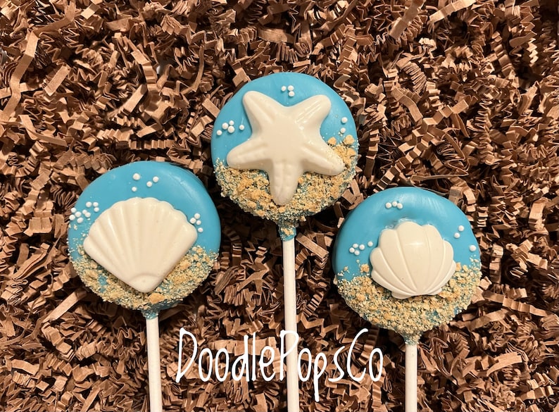 May include: Three blue cake pops with white seashell and starfish decorations. The pops are decorated with edible sand and white sprinkles. The pops are on white sticks, and the text "DoodlePopsCo" is visible. The background is brown.
