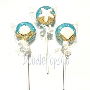 May include: Three blue-frosted lollipops with white seashell and starfish decorations, sprinkled with sand-colored edible granules. Each lollipop is wrapped in clear cellophane and tied with a white ribbon. The lollipops are on white sticks.