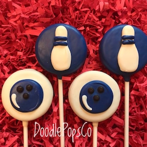 May include: Four round chocolate lollipops decorated with blue and white bowling-themed designs. Two lollipops feature a blue bowling pin with white stripes and a white bowling ball. The other two lollipops feature a blue bowling ball with white stripes and three black dots. The lollipops are on a red background. The text "Doodle PopsCo" is visible at the bottom of the image.