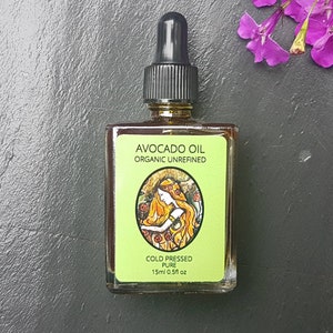 May include: A glass bottle of organic, cold-pressed avocado oil. The label is green and features a drawing of a woman with long hair. The label reads "Avocado Oil Organic Unrefined Cold Pressed Pure 15ml 0.5fl oz."