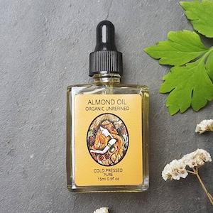 May include: A small glass bottle with a black dropper top filled with a clear liquid. The label is yellow with black text that reads "Almond Oil Organic Unrefined Cold Pressed Pure 15ml 0.5fl oz".
