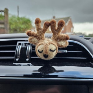 May include: A felted deer head car air vent clip. The deer has closed eyes, a black nose, and brown antlers. The deer is tan and brown in color. The clip is attached to a black car vent.