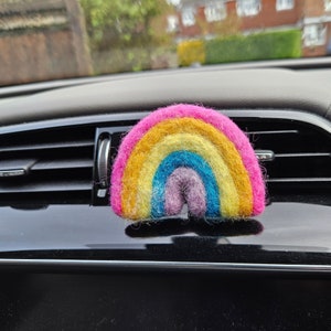 May include: A felted rainbow car air freshener in shades of pink, yellow, orange, blue, and purple. The rainbow is positioned on a black car dashboard, adding a touch of colour.