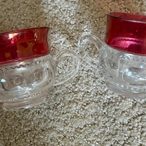 Peut inclure: Deux récipients en verre transparent avec des bords rouge foncé et des motifs texturés. L'un possède deux anses latérales, tandis que l'autre est un pichet avec une seule anse et un bec verseur. Les deux ont une base en verre pressé.