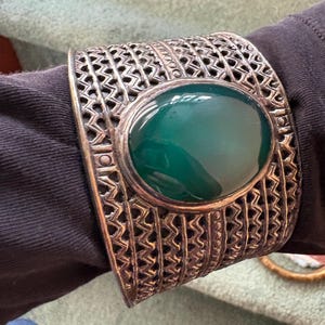 May include: A wide silver-toned cuff bracelet featuring an intricate openwork lattice pattern and a large, smooth, oval-shaped dark green stone set prominently in the center. The metalwork displays a textured, oxidized finish throughout.