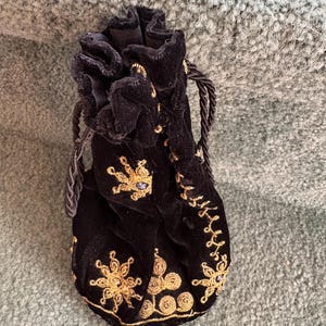 May include: A black velvet drawstring pouch featuring intricate gold thread embroidery in floral and swirling patterns, accented with small purple stones. The bag has a gathered top closure with matching black twisted cord drawstrings.