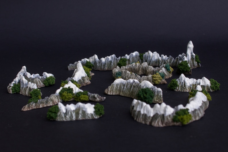 May include: A set of miniature mountain terrain pieces for tabletop gaming. The pieces are made of a light gray material with a textured surface and are painted with white snow and green foliage.