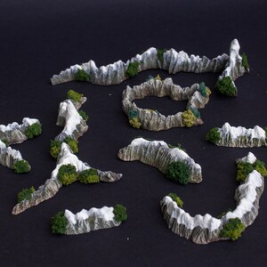 May include: A set of ten miniature mountain ranges with snow-capped peaks and green foliage. The mountains are made of a light gray material and are designed for tabletop gaming or model building.