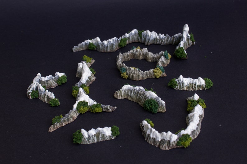 May include: A set of ten miniature mountain ranges with snow-capped peaks and green foliage. The mountains are made of a light gray material and are designed for tabletop gaming or model building.