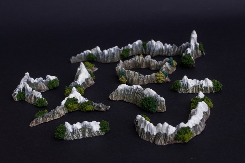 May include: A set of miniature mountain terrain pieces for tabletop gaming. The pieces are made of a light gray material with green accents and are designed to resemble snow-capped mountains.