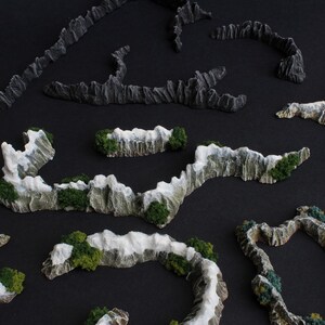 May include: A set of miniature mountain range terrain pieces for tabletop gaming. The pieces are made of a light gray material with white snow accents and green foliage.