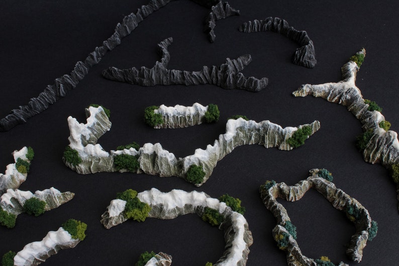 May include: A set of miniature mountain range terrain pieces for tabletop gaming. The pieces are made of a light gray material with white snow accents and green foliage.