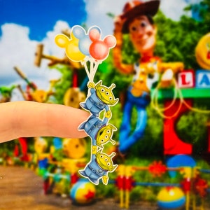 May include: Three green alien characters with three eyes each, wearing blue and white space suits, are holding onto each other and hanging from three balloons. The balloons are yellow, blue, and pink with a Mickey Mouse head design.