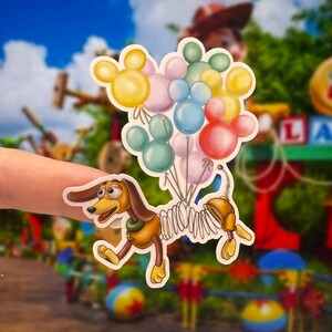 K&ouml;nnte beinhalten: Ein Aufkleber von Slinky Dog aus Toy Story, der einen Strau&szlig; bunter Ballons mit Mickey-Maus-Ohren h&auml;lt. Die Ballons sind rosa, gelb, blau, gr&uuml;n und rot.