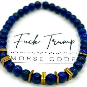 Puede incluir: Una pulsera de cuentas con cuentas esféricas azul oscuro y detalles rectangulares dorados. La pulsera tiene el texto "Fuck Trump" y "Morse Code" en escritura negra.