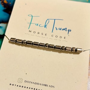 Peut inclure: Un bracelet en code Morse argenté épelant "Fuck Trump" sur une carte blanche. La carte porte le texte "Morse Code" sous la phrase et le nom de la boutique Etsy "DOTANDDASHBEADS". Le fond est un mélange de bleu et de vert.