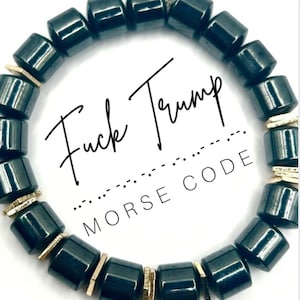 Op de afbeelding: Zwart kralenarmband met rechthoekige kralen en goudkleurige accenten. De armband heeft de tekst "Fuck Trump" en "Morse Code" in een witte cirkel. De kralen zijn donker.