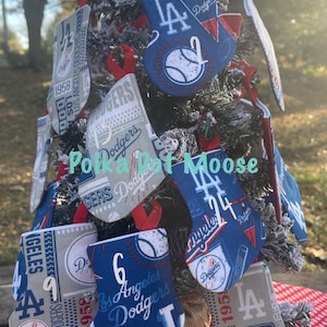 May include: A decorated Christmas tree featuring multiple Los Angeles Dodgers-themed mini stockings. The stockings are in blue, white, and gray, with the Dodgers logo, baseballs, and team-related text. Red ribbon loops are attached for hanging. The tree is set on a red and white polka dot surface.