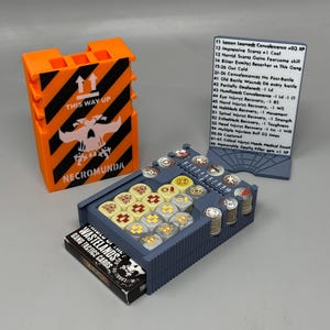 May include: An orange and black container with the text "THIS WAY UP" and "NECROMUNDA" next to a blue game organizer filled with game pieces and a card deck labeled "WASTELANDS GANG TACTICS CARDS". A white card with game rules is also present.