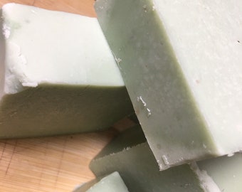 May include: Four bars of pale green soap stacked on a wooden surface. The soap has a slightly textured surface.