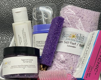 May include: A white wicker basket filled with lavender-themed bath and foot care products. The products include a jar of lavender foot scrub, a bag of lavender bath salts, a lavender foot cream, a purple foot file, a pink foot brush, and a set of manicure tools.