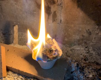 May include: A small, round, gray fire starter with a brown, fibrous material inside is burning with a bright yellow flame in a fireplace.