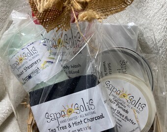 May include: A gift basket filled with natural bath and body products from Sapo de Solis. The basket includes a bar of tea tree and mint charcoal soap, a jar of shea butter hair pomade, a bottle of peppermint shower steamers, and a bottle of body wash.