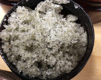May include: A close-up of a black container filled with a green and white sugar scrub. The scrub is made with sugar, oil, and herbs.