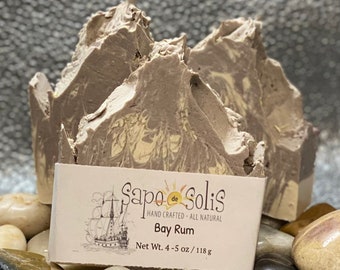 May include: Two bars of handmade Bay Rum soap, with a marbled brown and white design, sitting on top of gray stones. The soap is labeled "Sapo de Solis" and "Hand Crafted - All Natural".