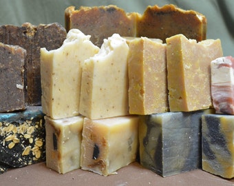 May include: Assortment of handmade soap bars in various colors and patterns, including brown, beige, and gray. The soaps are arranged in a pyramid shape on a brown surface.