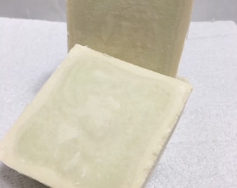 May include: Two rectangular bars of handmade soap. The soap has a creamy, off-white color with a subtle green hue in the center. The bars are sitting on a white, textured surface.