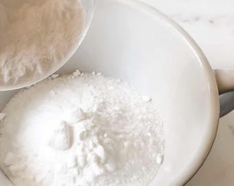 May include: A white ceramic bowl being filled with a white powder from a clear glass container. The powder is being poured into the bowl, creating a cloud of white. The bowl is on a white surface.