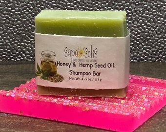 May include: A green bar of soap with a white label that reads "Sapo Solis Hand Crafted - All Natural Honey & Hemp Seed Oil Shampoo Bar Net Wt. 4-5 oz/113g". The soap is sitting on a pink and white glittery surface.