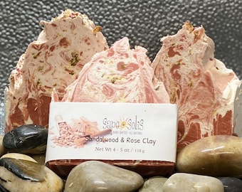 May include: Three bars of handmade sandalwood and rose clay soap. The soap is a light pink color with swirls of brown and white. The soap is sitting on a bed of small gray rocks. The label on the soap reads "Sapo Solis Hand Crafted All Natural Sandalwood & Rose Clay Net Wt 4-5 oz / 118 g"
