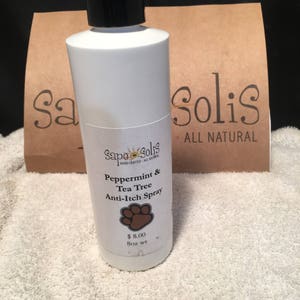 May include: A white plastic bottle with a black spray nozzle and a label that reads "Sapo Solis Hand Crafted - All Natural Peppermint & Tea Tree Anti-Itch Spray". The bottle is sitting on a brown paper bag with the words "Solis All Natural" printed on it.