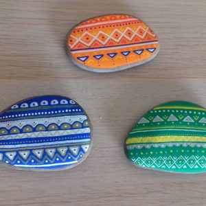 Peut inclure: Trois pierres peintes avec des motifs géométriques. Les pierres sont peintes en bleu, vert et orange avec des accents blancs.