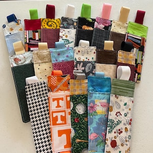 Puede incluir: Una colección de marcapáginas de tela hechos a mano con varios patrones y colores. Los diseños incluyen estampados florales, pata de gallo y logotipos de la Universidad de Tennessee. Cada marcapáginas tiene una cinta a juego, con colores como verde, rosa y azul.