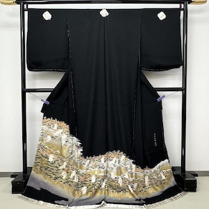 May include: A black kimono with a landscape design at the hem, displayed on a black rack. The landscape features trees and mountains in shades of gold, white, and gray. The kimono has white floral accents near the shoulders.
