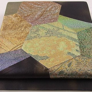 Può includere: Tessuto piegato con un design patchwork. Lo strato superiore presenta forme geometriche in oro, verde, viola e blu. Lo strato inferiore è nero tinta unita. Il tessuto ha un aspetto strutturato e fantasia.